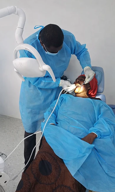 Dentist working on a female patient in a dental chair. They are both wearing light blue protective covering.